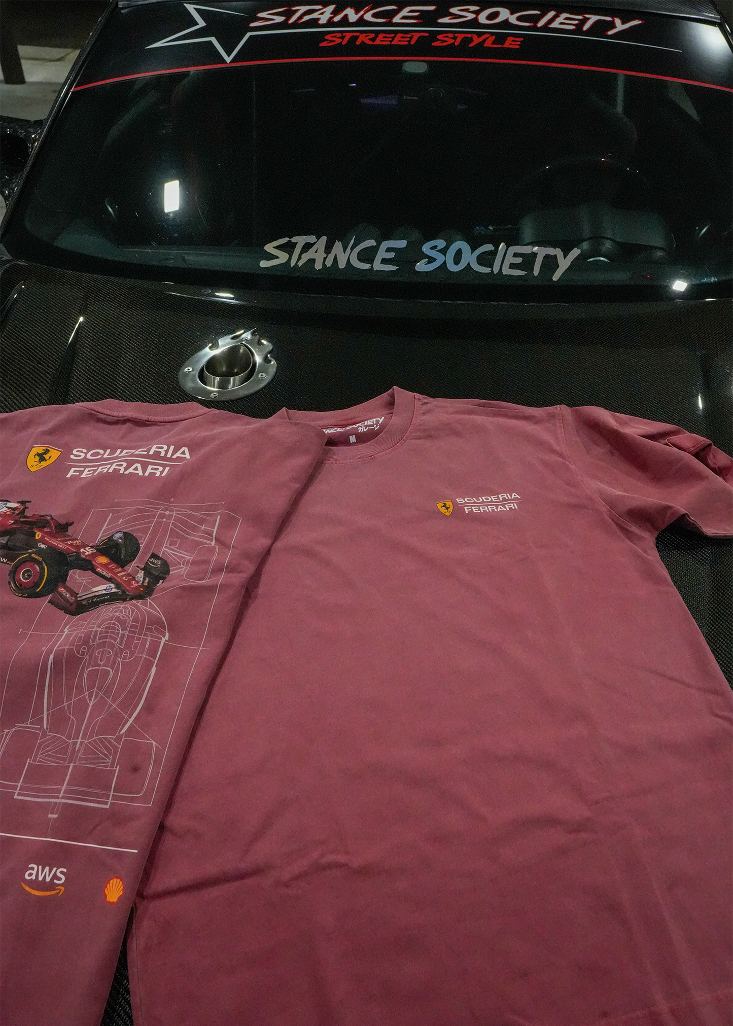 A pair of vintage red short-sleeve t-shirts featuring Ferrari graphics, laid flat on the hood of a black car. The left shirt displays a large graphic of a red Ferrari F1 car and technical line drawings, while the right shirt has a small yellow and red Ferrari logo on the left chest.