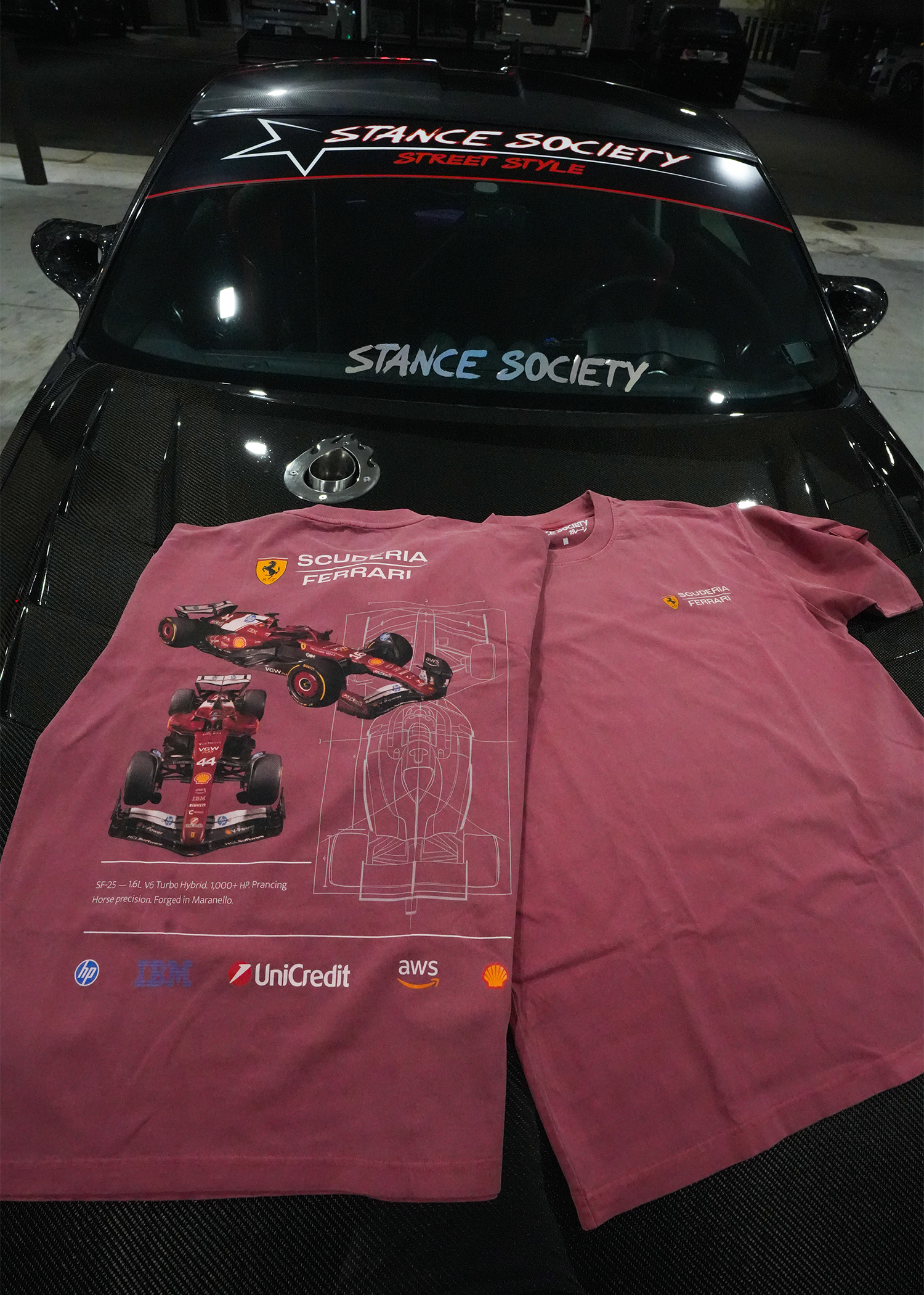 A pair of Vintage Red t-shirts featuring a Ferrari Scuderia Ferrari F1 design, laid flat on the hood of a black car. The left shirt shows a detailed graphic of a red Formula 1 car, while the right shirt shows the plain back side.